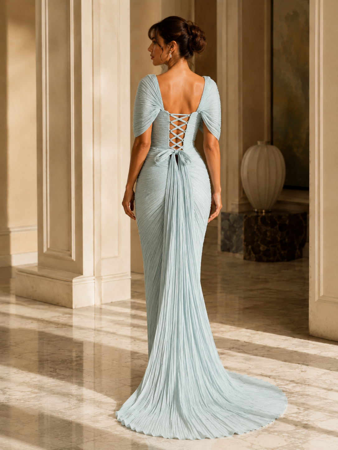 A woman wearing a light blue pleated evening gown, photographed from the back in an elegant indoor hallway with columns and a marble floor.