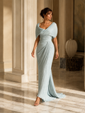 A woman models a floor-length light blue /green pleated gown with a draped one-shoulder design in an elegant indoor setting with warm sunlight and cream columns.