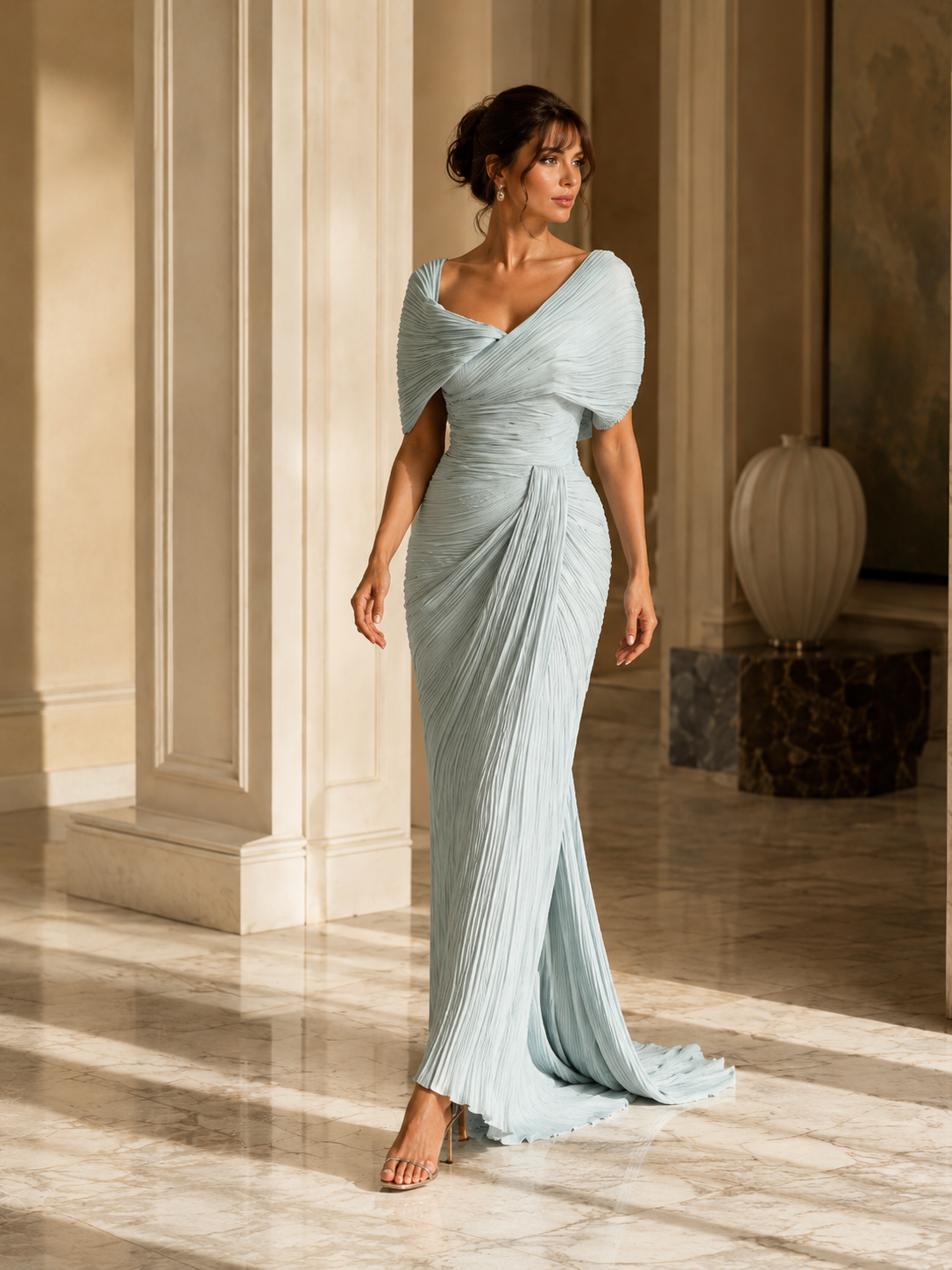 A woman models a floor-length light blue /green pleated gown with a draped one-shoulder design in an elegant indoor setting with warm sunlight and cream columns.