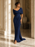 A woman models a floor-length navy blue off-the-shoulder gown with draped short sleeves and a gathered, pleated skirt in a sunlit indoor hallway with marble columns and floors.