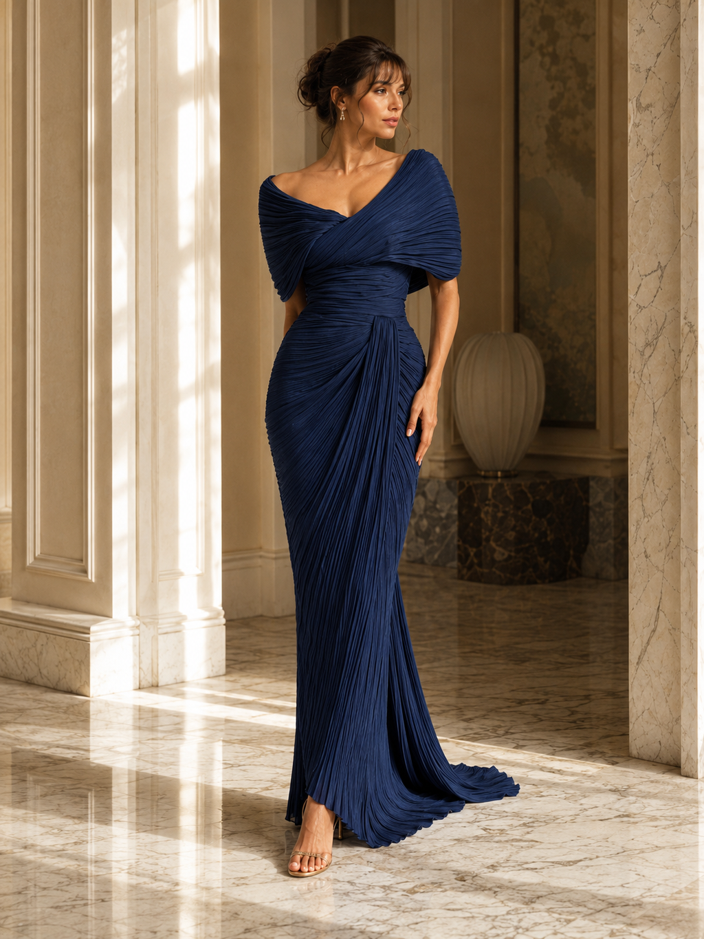 A woman models a floor-length navy blue off-the-shoulder gown with draped short sleeves and a gathered, pleated skirt in a sunlit indoor hallway with marble columns and floors.
