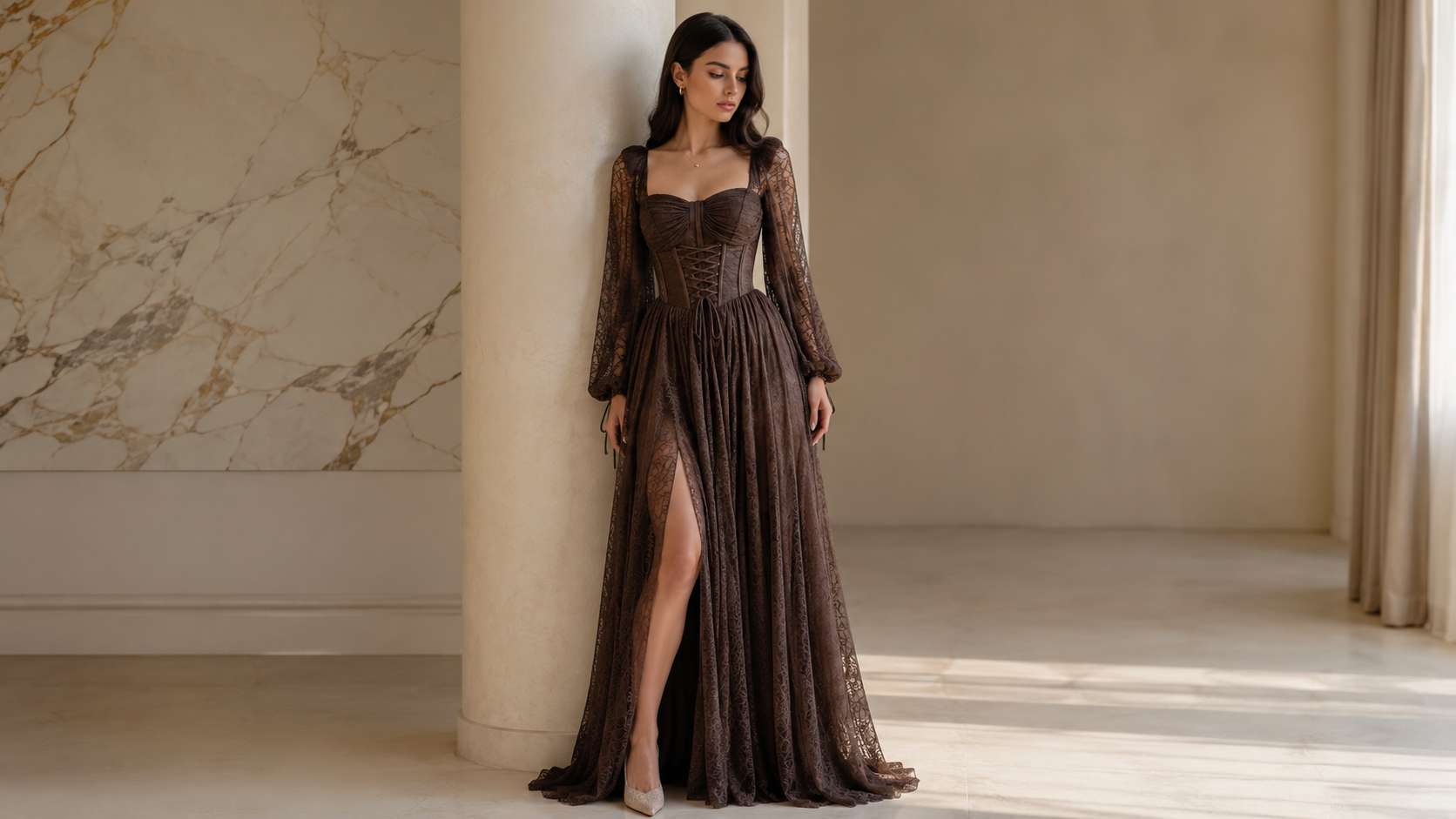 A woman wearing a floor-length brown lace gown with long sheer sleeves and a high slit, standing indoors beside a large cream column in a softly lit room.

