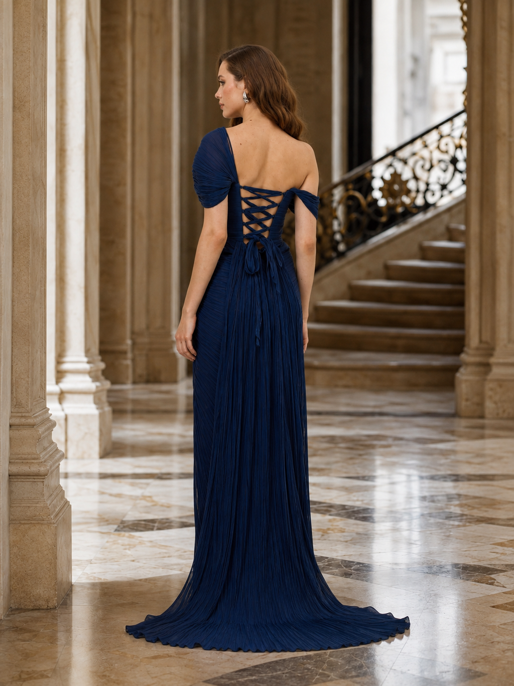 A woman wearing a floor-length navy blue evening gown with an off-the-shoulder neckline, lace-up open back, and long pleated train, photographed in a grand indoor hallway with columns and a staircase.

