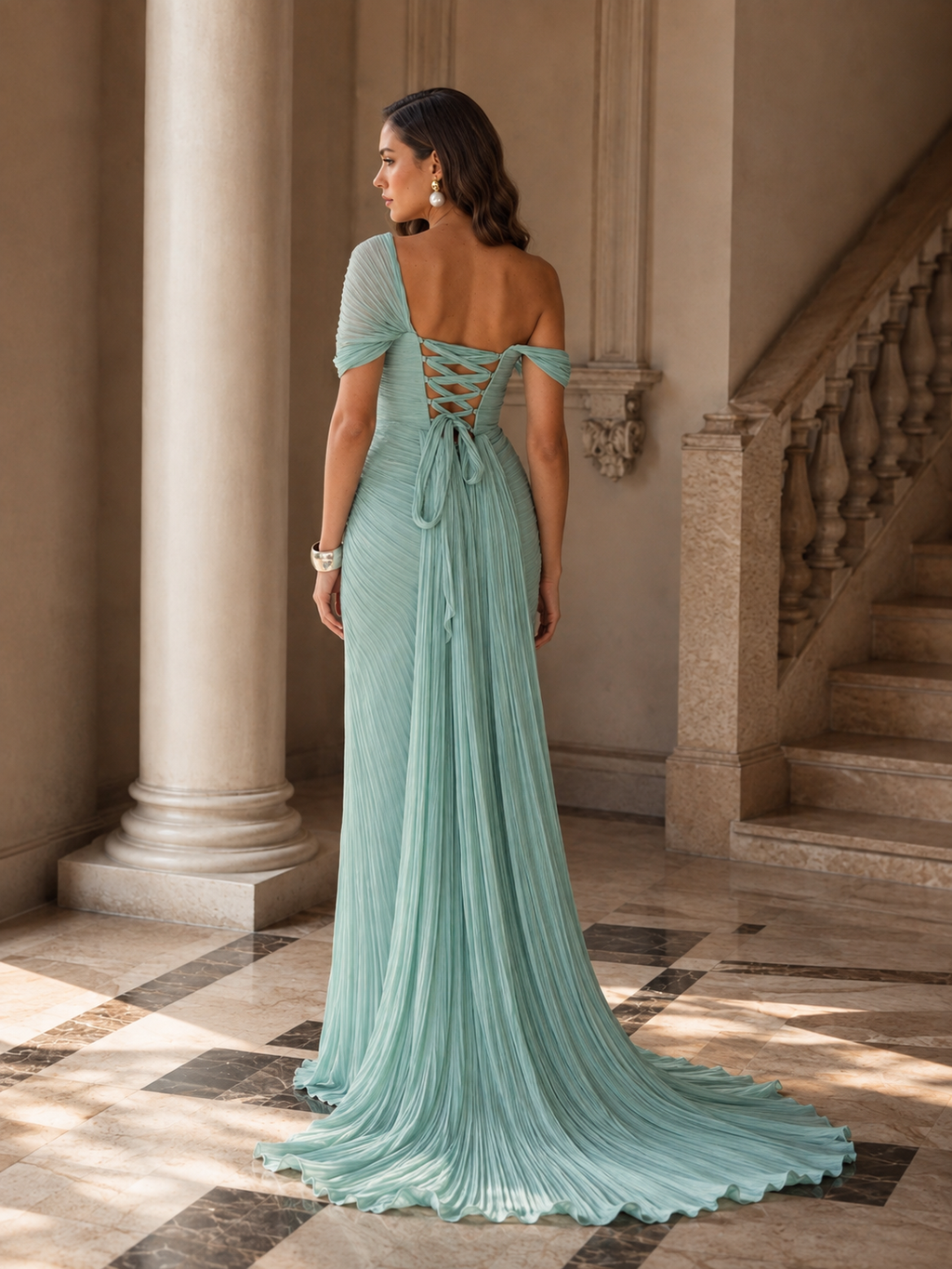 A woman wearing a flowing mint green off-the-shoulder evening gown with an open lace-up back, photographed in a grand interior hallway with columns and a staircase.

